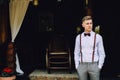 A handsome young man in a shirt, a bow tie, trousers and suspenders stands on the veranda of the house.