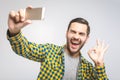 Handsome young man in shirt holding camera and making selfie and smiling while standing against grey background. Ok Royalty Free Stock Photo