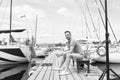 Handsome young man sitting on bench in dock of the bay between boats. Serious boat man on pier looking away Royalty Free Stock Photo