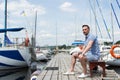 Handsome young man sitting on bench on pier between moored boats. Young businessman on vacation on pier