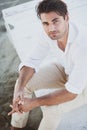 Handsome young man sitting looking up. He wears a white shirt and brown trousers.