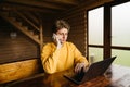 Handsome young man sitting in a wooden room in the country with a laptop and talking on the phone. Guy works on vacation in a Royalty Free Stock Photo
