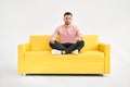 Handsome young man sitting in yoga lotus pose and meditating on comfortable yellow sofa Royalty Free Stock Photo