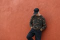 Handsome young man in stylish black cap in military fashionable shirt in trendy jeans rests standing near a vintage red wall Royalty Free Stock Photo