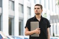 Handsome young man in stylish casual clothes standing on the street with a laptop in his hand, looking away, on the background of Royalty Free Stock Photo