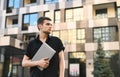 Handsome young man in stylish casual clothes standing on the street with a laptop in his hand, looking away, on the background of Royalty Free Stock Photo