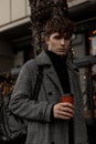 Handsome young man in a stylish plaid coat with a fashionable backpack stands with a cup of delicious coffee on the street.