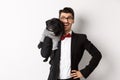 Handsome young man in suit and glasses holding cute black pug dog on shoulder, smiling happy at camera, wearing party Royalty Free Stock Photo