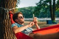 Handsome young man in sunglasses lying at sunset in a hammock and using a smartphone. The guy is resting in a hammock in the park