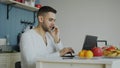 Handsome young man talking phone and using laptop computer sitting in the kitchen after breakfast in the morning Royalty Free Stock Photo