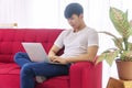 Man using laptop for working online activities while sitting on red couch at home. Freelance guy typing notebook