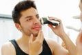 Handsome young man wearing black singlet top looking in mirror, using electric shaving machine during morning routine