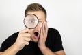 Handsome young man wearing black t-shirt holding magnifying glass near his eye looks surprised isolated white background Royalty Free Stock Photo