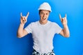 Handsome young man wearing builder uniform and hardhat shouting with crazy expression doing rock symbol with hands up