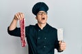 Handsome young man wearing chef uniform holding bacon and knife angry and mad screaming frustrated and furious, shouting with