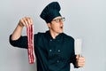 Handsome young man wearing chef uniform holding bacon and knife angry and mad screaming frustrated and furious, shouting with Royalty Free Stock Photo