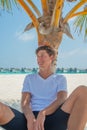 Handsome young man wearing white t-shirt and black shorts relaxing under palm tree at tropical sandy beach at island luxury resort Royalty Free Stock Photo