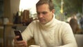 Handsome young man in a white sweater using a smartphone, sitting in a cafe in the city on an against a window Royalty Free Stock Photo