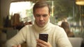 Handsome young man in a white sweater using a smartphone, sitting in a cafe in the city on an against a window Royalty Free Stock Photo