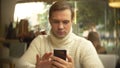 Handsome young man in a white sweater using a smartphone, sitting in a cafe in the city on an against a window Royalty Free Stock Photo