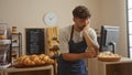 Handsome young man working in a bakery shop looking thoughtful with fresh pastries on display and a chalkboard menu in the Royalty Free Stock Photo