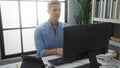 Handsome young man working at a computer in an office with bookshelves and a large window showing a bright, modern interior Royalty Free Stock Photo