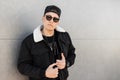 Handsome young men hipster in a stylish black jacket in a fashionable cap in dark trendy sunglasses in a black t-shirt stands Royalty Free Stock Photo