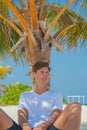 Handsome young model sexy man in stylish clothes relaxing under palm tree at tropical sandy beach at island luxury resort Royalty Free Stock Photo