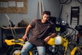 A handsome young motorcyclist in jeans and a t-shirt poses for a photo sitting on a bike in his garage