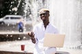 Handsome young pensive Afro American businessman is using a laptop while sitting outdoors in summer street near fountain at fresh Royalty Free Stock Photo