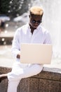 Handsome young pensive Afro American businessman is using a laptop while sitting outdoors in summer street near fountain at fresh Royalty Free Stock Photo