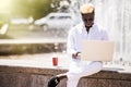 Handsome young pensive Afro American businessman is using a laptop while sitting outdoors in summer street near fountain at fresh Royalty Free Stock Photo