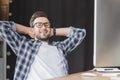 handsome young programmer in eyeglasses smiling at camera while sitting with hands behind head