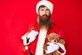 Handsome young red head man with long beard wearing santa claus costume holding teddy bear depressed and worry for distress, Royalty Free Stock Photo