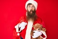 Handsome young red head man with long beard wearing santa claus costume holding teddy bear making fish face with mouth and