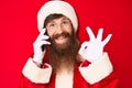 Handsome young red head man with long beard wearing santa claus costume talking on the phone doing ok sign with fingers, smiling Royalty Free Stock Photo