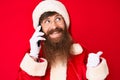 Handsome young red head man with long beard wearing santa claus costume talking on the phone pointing thumb up to the side smiling Royalty Free Stock Photo