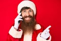 Handsome young red head man with long beard wearing santa claus costume talking on the phone smiling happy pointing with hand and Royalty Free Stock Photo