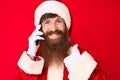 Handsome young red head man with long beard wearing santa claus costume talking on the phone smiling happy and positive, thumb up Royalty Free Stock Photo