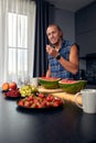 Handsome young sporty smiling man in the kitchen is preparing vegan healthy fruits salad and smoothie in a good mood Royalty Free Stock Photo