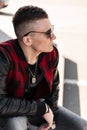 Handsome young stylish hipster man in sunglasses in trendy clothes is sitting on wooden steps in the city on a warm sunny day. Royalty Free Stock Photo