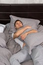 Handsome young tired man in pajama sleeping alone without blanket and hugging a pillow in stylish bed