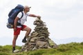 Handsome young tourist man with backpack making pyramid pile of stones on lit by bright summer sun green grass against light blue