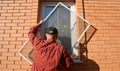 A handyman is installing anti-insect mosquito net, screen, fly and bugs protection on a plastic window of a brick house to keep
