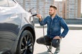 Hansome bearded guy sitting near his new modern electric car and holding plug of the charger, while car is charging at