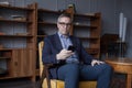 Hansome elderly businessman in blue suit and glasses working and looking on his mobile phone on office wall with boockshelf