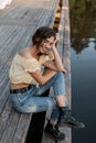 Happiness lonely curly woman with smile in summer fashionable clothes with blouse, jeans and boots sits and rest on a wooden pier