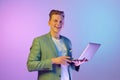 Happiness. Successful project. Young handsome smiling man in stylish jacket posing with laptop over purple background in Royalty Free Stock Photo