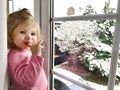 Happy adorable baby girl sitting near window and looking outside on snow on Christmas day or morning. Smiling healthy Royalty Free Stock Photo