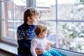 Happy adorable kid boy and cute baby girl sitting near window and looking outside on snow on Christmas day or morning Royalty Free Stock Photo